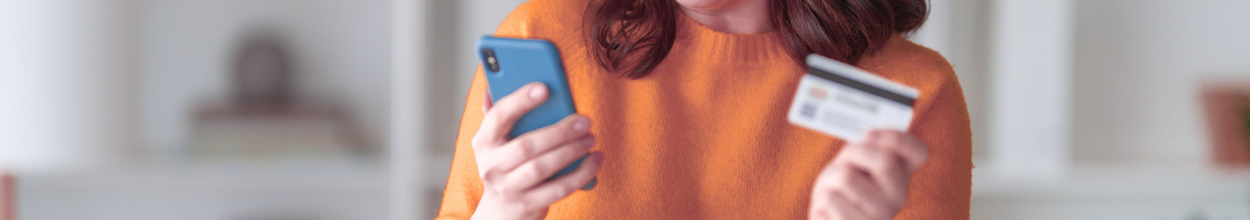 Primo piano di una persona in maglione arancione che tiene uno smartphone e una carta di credito per un acquisto online.