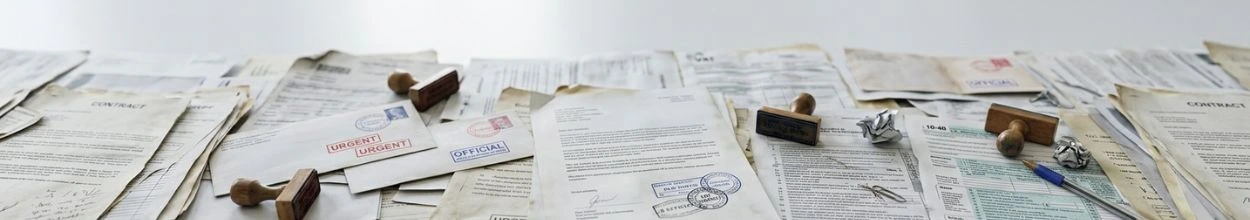 Una scrivania interamente ricoperta da una distesa di documenti cartacei disordinati, contratti, buste da lettera con timbri rossi "URGENT" e diversi timbri datari in legno sparsi sui fogli.
