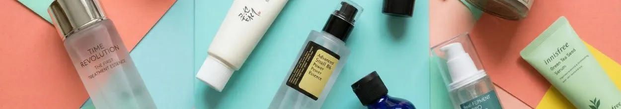 Una vista dall'alto di un assortimento di popolari prodotti per la cura della pelle coreani (K-beauty) disposti su uno sfondo geometrico di cartoncini colorati color pesca, acquamarina e giallo. Sono visibili flaconi di essenze, sieri e tubetti di crema