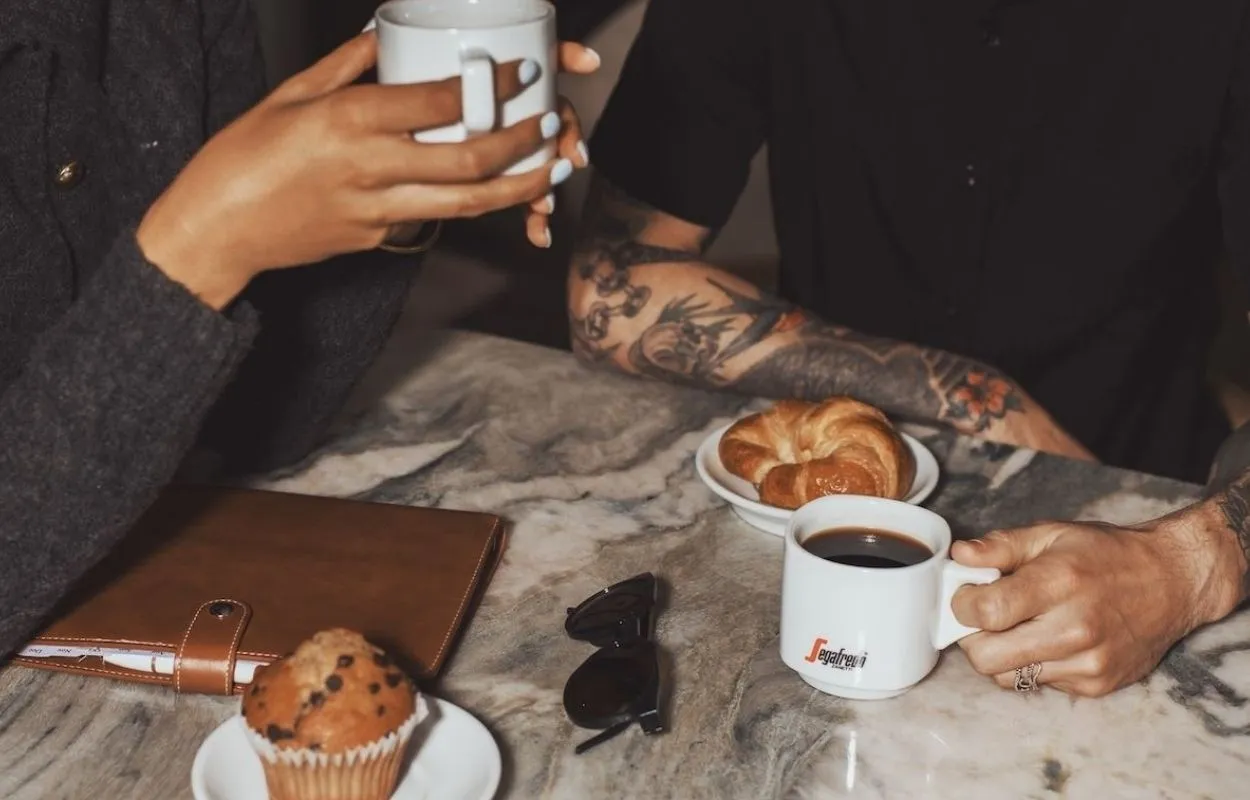Un primo piano di due persone (una con braccia tatuate) che bevono caffè Segafredo da tazze bianche. Sul tavolo di marmo ci sono un croissant e un muffin.