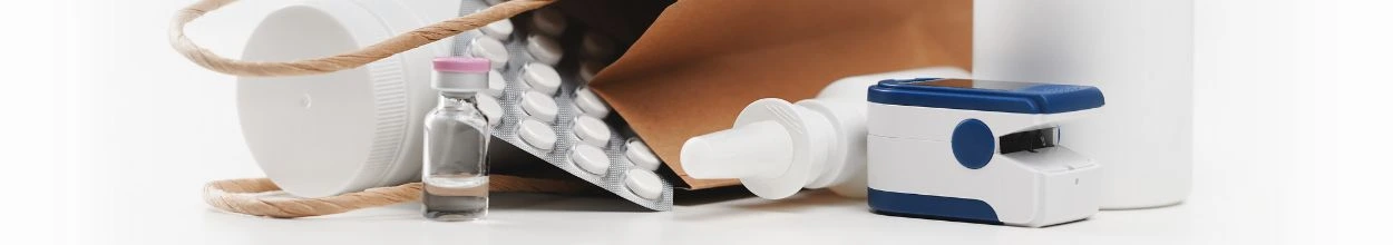 Sporta di carta con prodotti farmaceutici: saturimetro, flacone di gocce oculari, bottiglino in vetro, blister di pastiglie e barattolo per compresse.